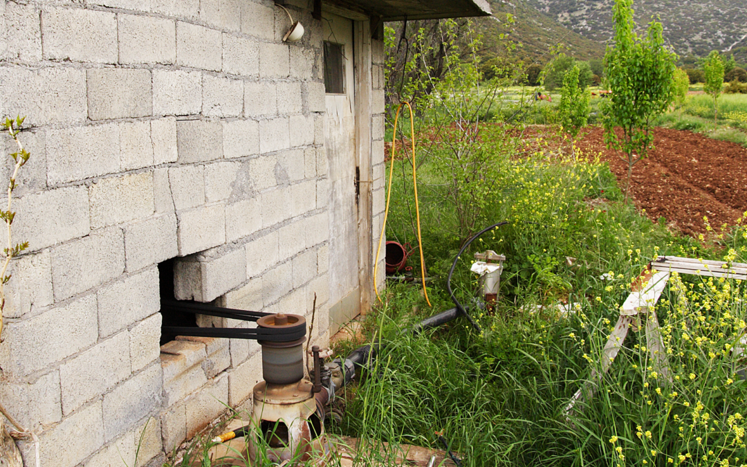 Weathered concrete pump house and belt-driven well pump beside a tilled farm plot with hills in the background